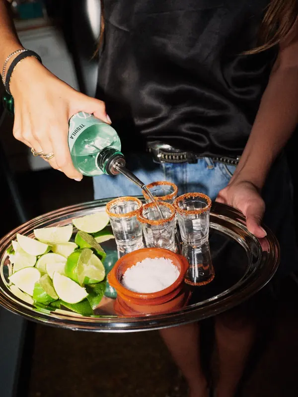 Pouring De Nada Blanco into four tajín-rimmed shot glasses on a silver tray with limes and salt