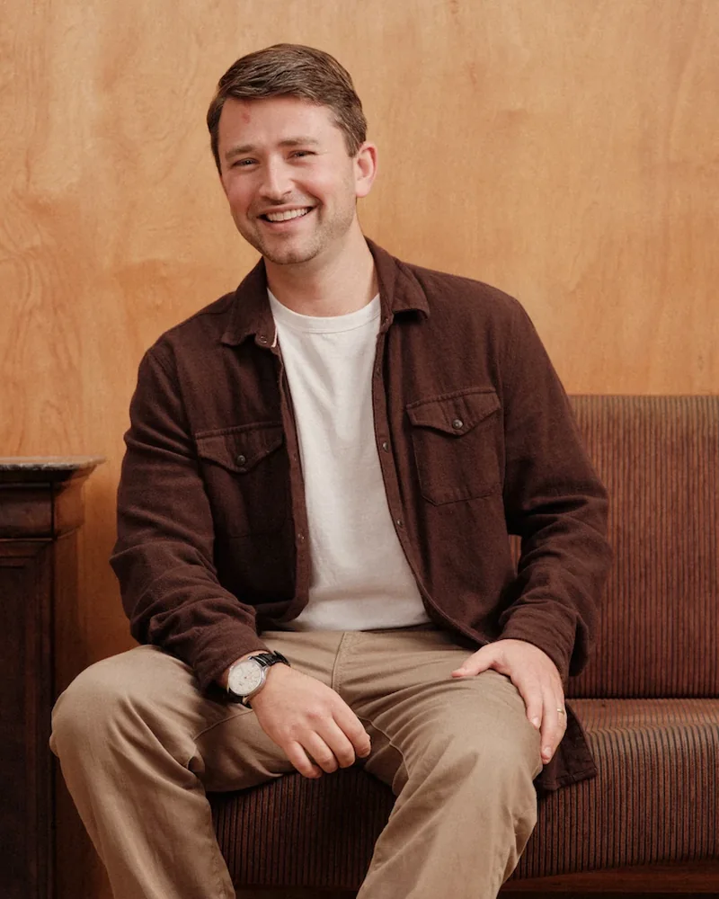 Adam Millman on a brown sofa against a wood-panelled wall, smiling