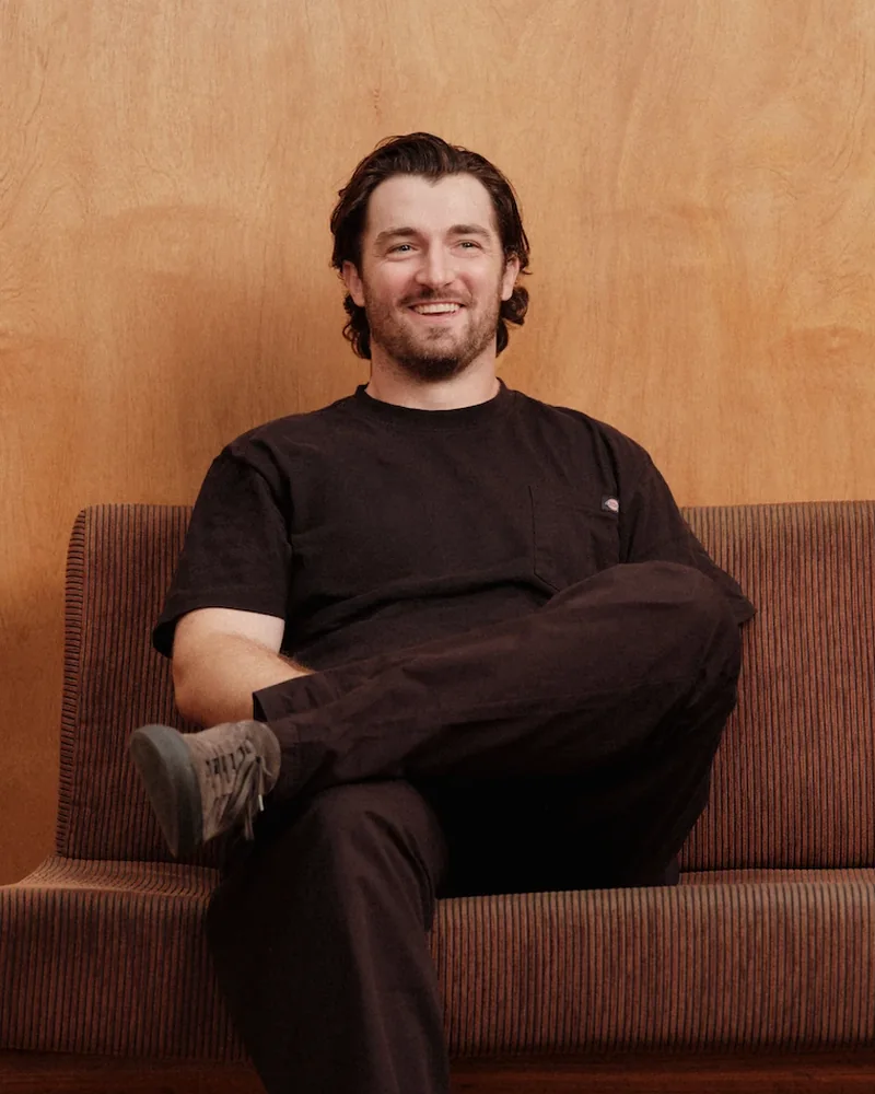 Danny Neeson on a brown sofa against a wood-panelled wall, legs crossed, smiling