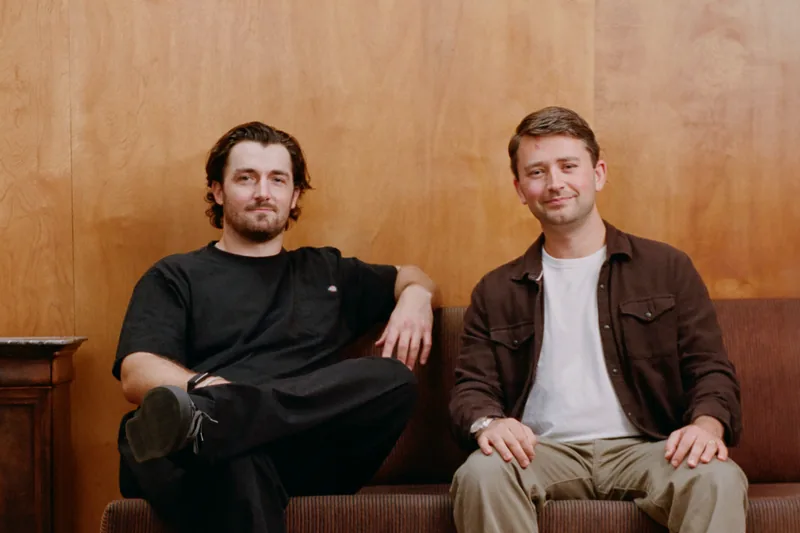 Danny Neeson and Adam Millman sitting on a brown sofa against a wood-panelled wall, shot on film